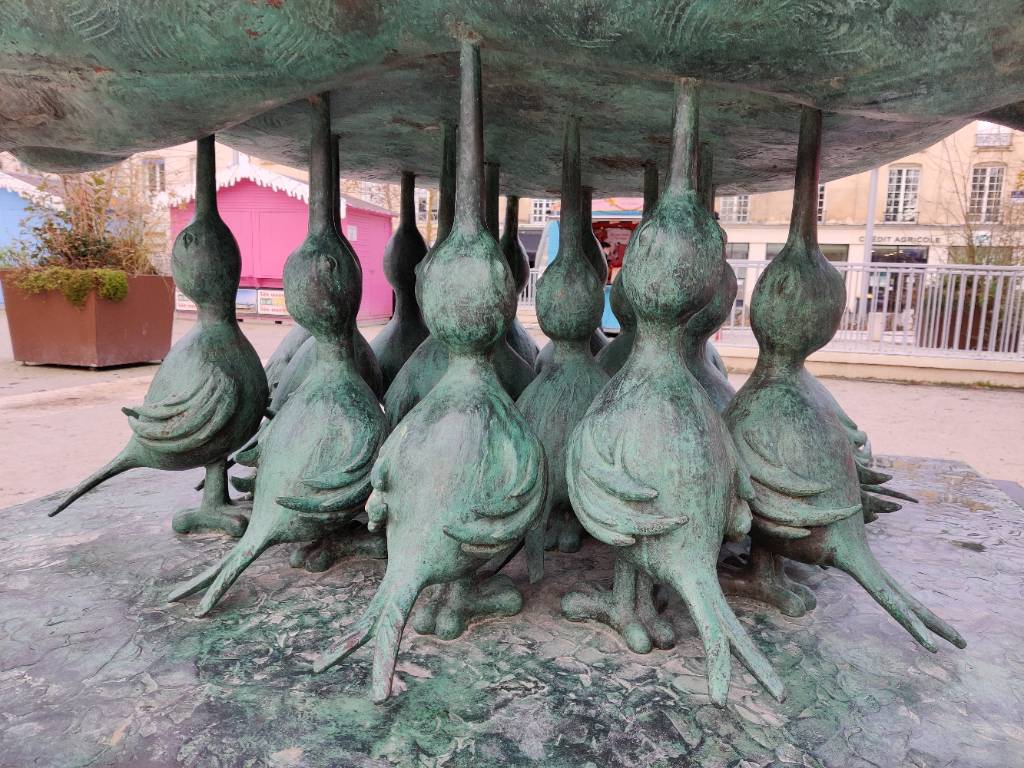 Détail d'une sculpture en bronse, une douzaine d'oiseaux tête levée, la pointe du bec portant une masse