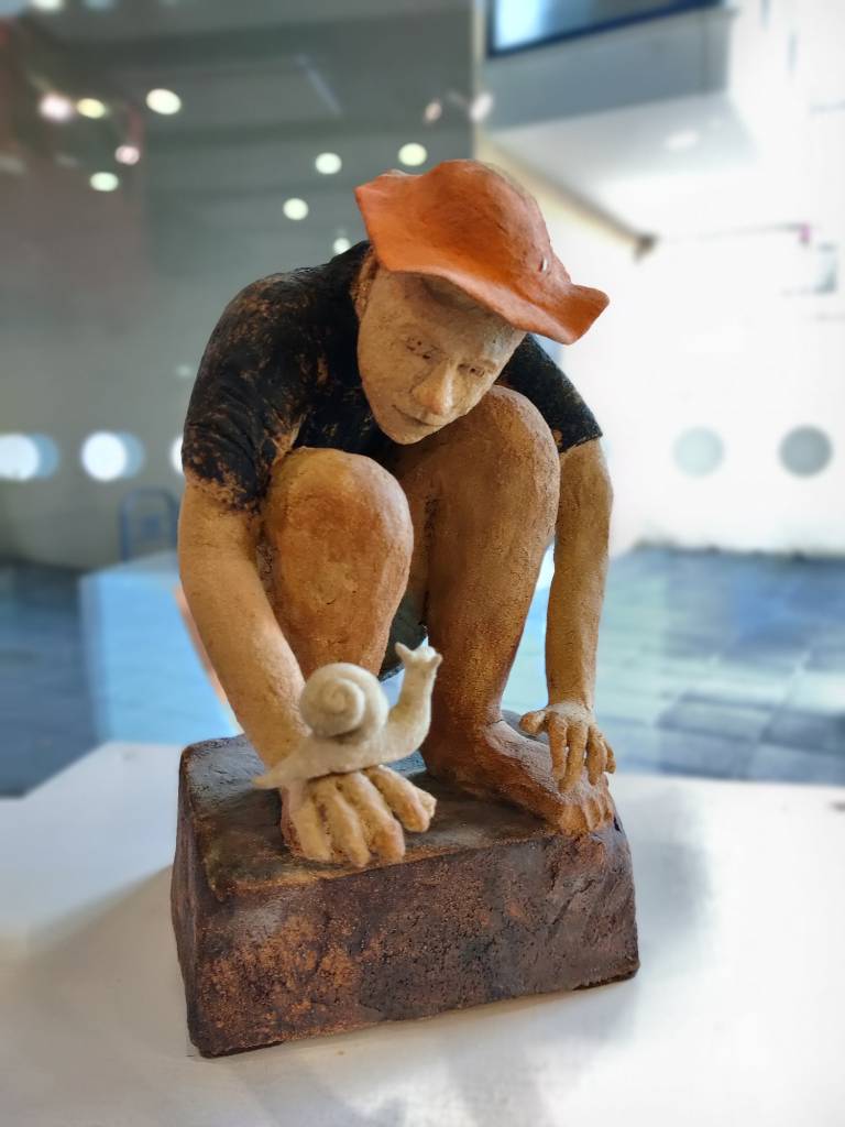 Sculpture en argile représentant un homme accroupi regardant un escargot sur sa main, le tout dans un style assez BD