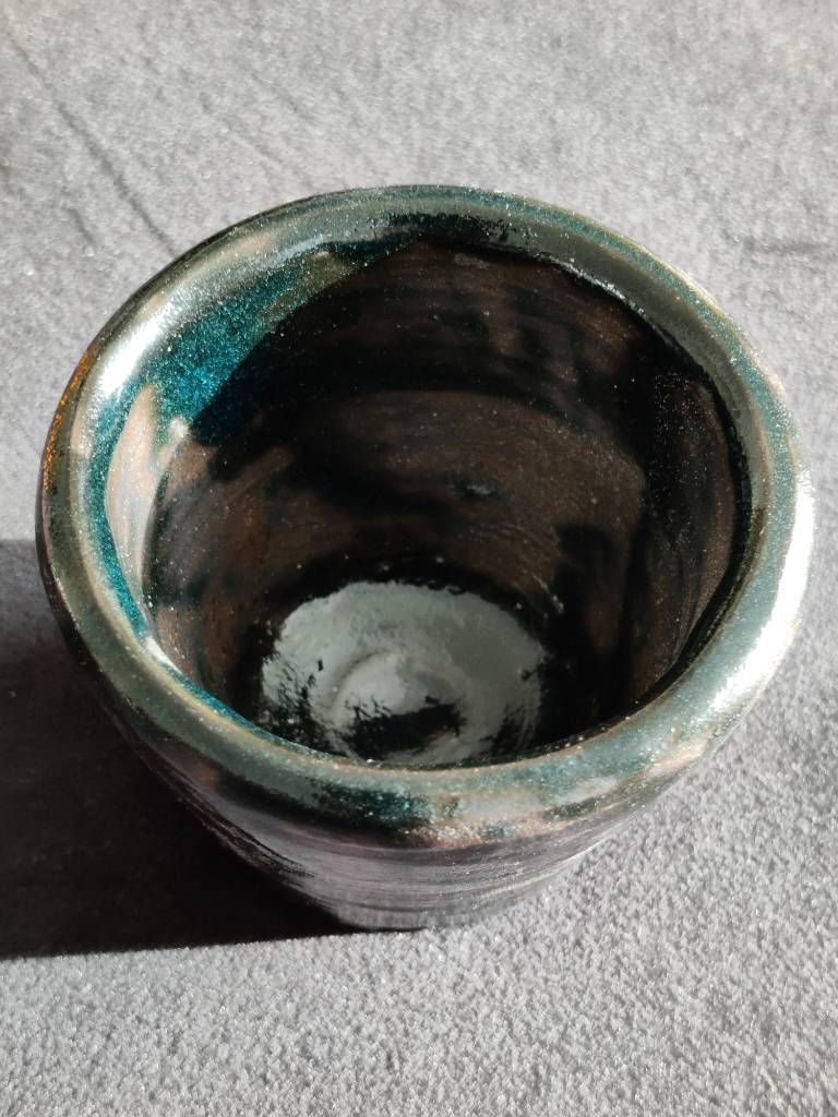 Détail de l'intérieur d'un petit pot en argile recouvert d'émail vert/noir (avec des reflets bleus sur la photo) posé de façon très irrégulière.
