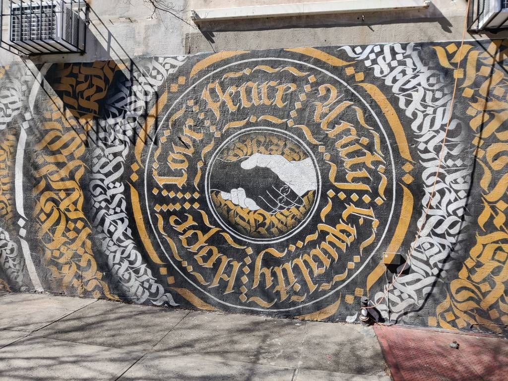 Graffiti représentant une poignée de main entre une main noire et une blanche au milieu d'un cercle de calligraphie latine ("Love, Peace, Unity, Equality, Hope") et calligraphie ornementale