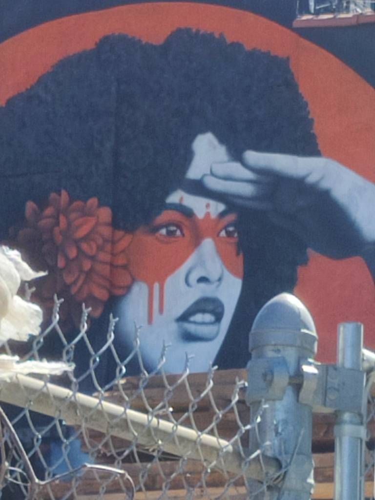 Détail d'un graffiti qui était caché où l'on voit une femme avec une coupe afro, une fleur rouge dans les cheveux et de la peinture rouge autour des yeux regarder au loin sa main sur le front