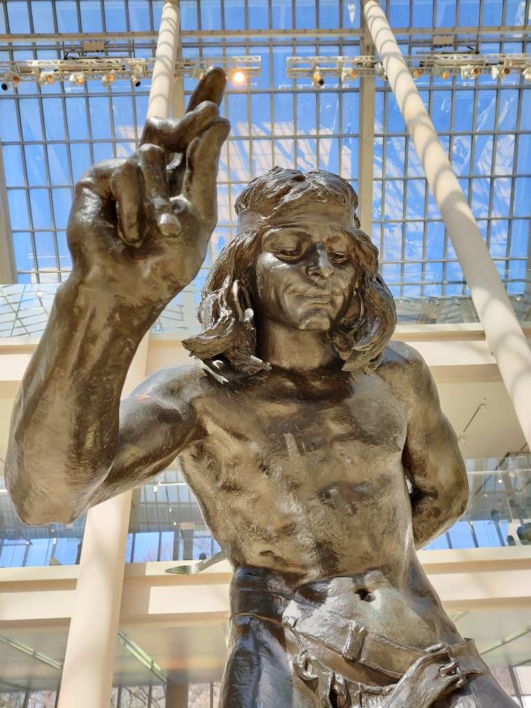 Sculpture vue du bas représentant un homme levant le doigt (il est en fait en train d'éduquer un ourson)