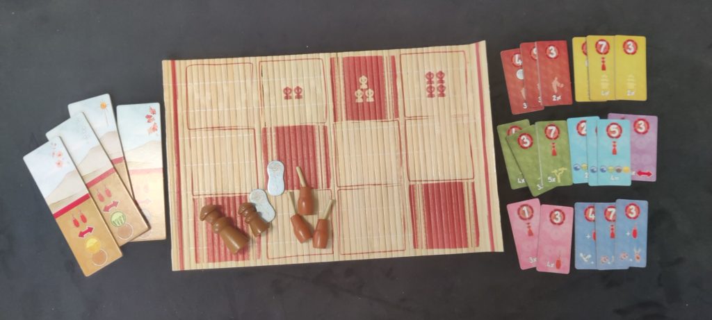 Divers éléments : des cartons avec une illustration et des outils, un plateau de jeu en nappe de bambou, des pions, des cartons de couleurs différentes avec des points