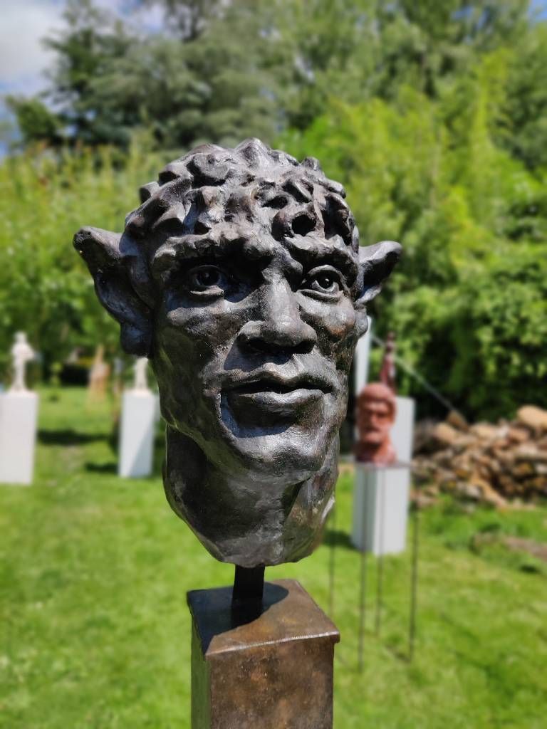 Une sculpture de tête en bronze dans un jardin. À l'arrière, on devine d'autres sculptures sur leur socle