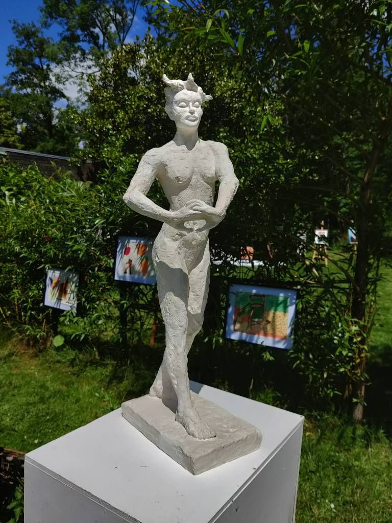 Une sculpture d'une femme nue debout les mains jointes devant elle dans un jardin. À l'arrière, des tableaux sont accrochés sur une haie