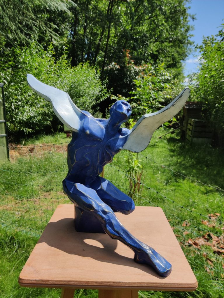 Une sculpture bleue, anguleuse, représentant un homme dont les bras sont des ailes, le visage tourné vers le ciel