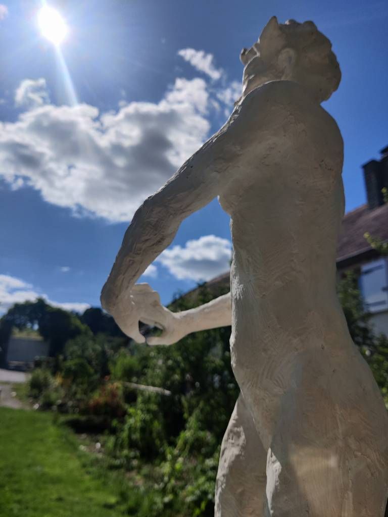 Détail d'une sculpture d'une femme joignant ses mains devant elle au premier plan et sur le côté. Le reste laisse voir un bout de jardin, une maison, quelques nuages et le soleil