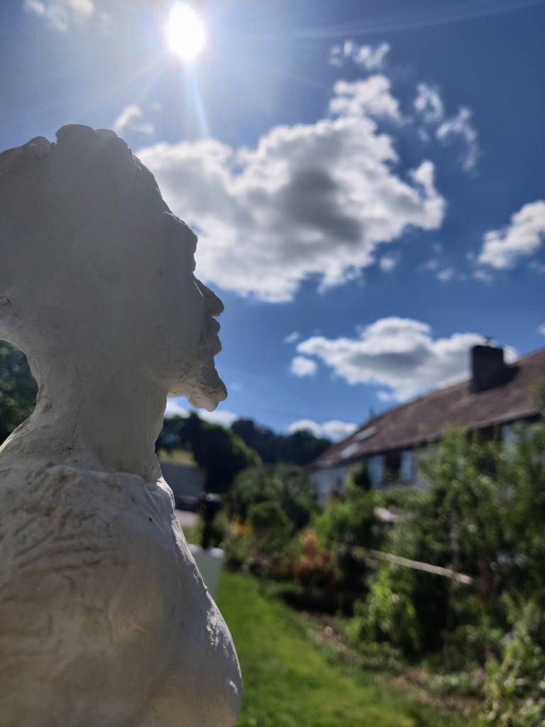 Détail d'une sculpture d'un homme de profil au premier plan et sur le côté. Le reste laisse voir un bout de jardin, une maison, quelques nuages et le soleil